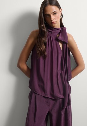 Donna con lunghi capelli scuri indossa una camicetta viola senza maniche con nodo al collo e pantaloni larghi coordinati, in piedi contro un muro chiaro e semplice.