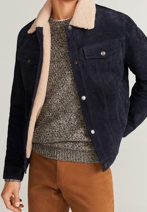 Man in een marineblauwe suède jas met beige shearlingkraag, gedragen over een grijze gebreide trui en bruine broek, staand met de hand langs het lichaam.