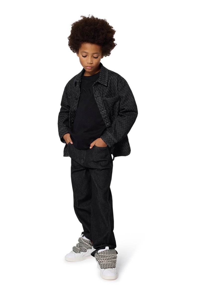 Veste en denim noire à motifs, t-shirt noir, pantalon en denim foncé et baskets blanches avec des accents tricotés noir et blanc. Les chaussures ont des détails texturés.