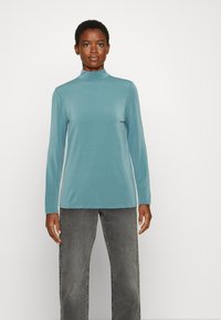 Top de manga longa azul claro com gola alta, feito de um tecido suave. Apresenta um corte descontraído, combinado com calças cinzentas.