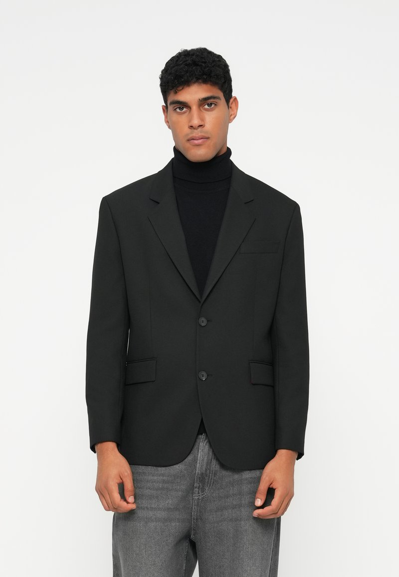 Blazer preto com dois botões, lapelas recortadas e dois bolsos frontais. Usado sobre um suéter de gola alta preto, combinado com jeans cinza. Textura suave.