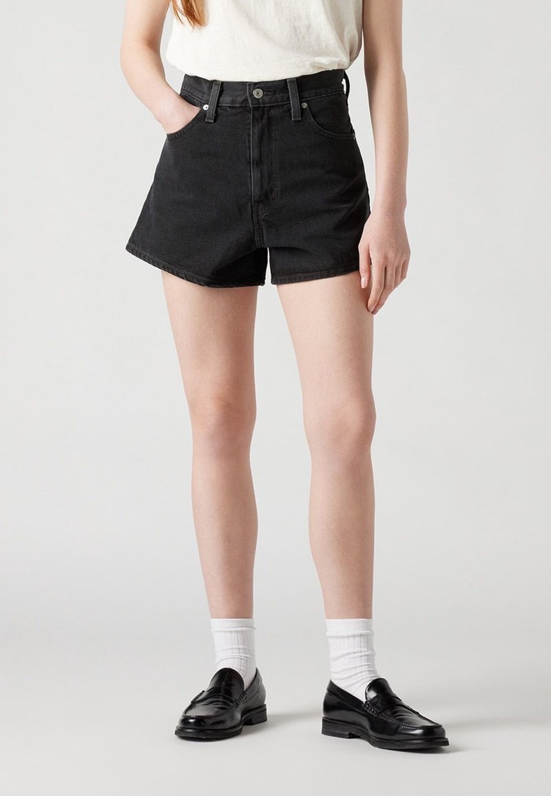 Jeune personne portant un short en jean noir, des chaussettes blanches, des mocassins noirs et un haut blanc sans manches, debout devant un fond clair uni.