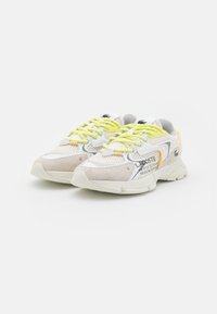 Lacoste sneakers in white and beige mesh with suede accents. Features bright yellow laces, logo detail, and a textured rubber sole.
