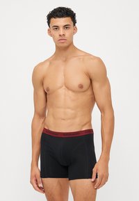 Schwarze Boxershorts aus dehnbarem Stoff mit einem dunkelroten Gummibund, auf dem das Logo "BJÖRN BORG" in Schwarz angezeigt wird.