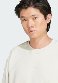 Light beige sweatshirt made of soft fabric, featuring a ribbed crew neckline and subtle embroidered logo on the chest. Smooth texture, relaxed fit.