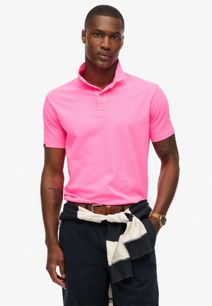 Polo à manches courtes en coton rose vif, avec un col montant et une patte de boutonnage à trois boutons. Associé à un pantalon foncé et un léger pull noué.