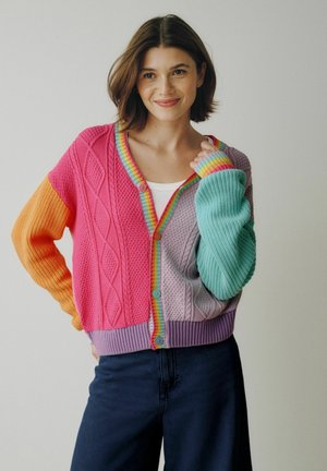 Noor naine, kellel on lühikesed pruunid juuksed, kannab värvilist kootud kardigani roosa, oranži, lilla ja türkiissiniste aladega, naeratades käsi krae lähedal.
