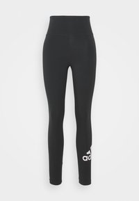 Černé legíny Adidas s vysokým pasem, přiléhavými nohavicemi a bílým logem Adidas na dolní části levé nohavice na jednobarevném pozadí.