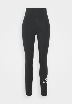 Leggings noires taille haute d'Adidas avec des jambes ajustées et le logo blanc d'Adidas sur la jambe inférieure gauche sur fond uni.