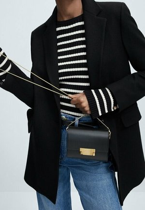 Persona che indossa un blazer nero, un maglione a righe bianche e nere, jeans blu, e tiene una piccola borsa nera con fibbia e tracolla dorate.
