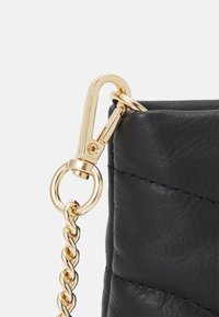 Angolo di borsa in pelle nera con una clip metallica dorata e un attacco a catena intrecciata dorata. Superficie testurizzata con cuciture visibili.