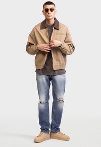 Beige bomber jacket with a brown corduroy collar, worn over a dark shirt; paired with distressed blue jeans and light brown sneakers.