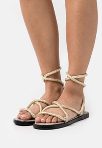 Infradito piatti con suola nera e dettagli in corda intrecciata beige. Design open-toe che mette in risalto il tono di pelle chiaro e una manicure delle unghie dei piedi ordinata.