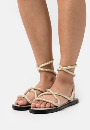 Sandales plates à brides avec une semelle noire et des détails en corde tressée beige. Le design ouvert met en valeur un teint de peau clair et un vernis à ongles soigné.