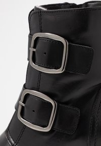 Černá kožená kotníčková bota se dvěma stříbrnými sponami, hladkým povrchem a detailem šití. Design zahrnuje zakulacený horní okraj.