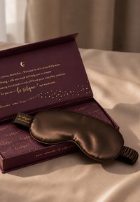 Maschera per occhi marrone setosa che riposa in una scatola viola con testo dorato che promuove il relax e la cura delicata della pelle intorno agli occhi.