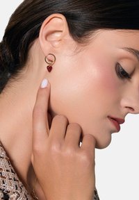 Złoty kolczyk w kształcie koła z małym czerwonym serduszkiem. Projekt jest delikatny, z gładkim wykończeniem i prostą okuciem.