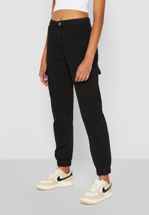 Femme portant un pantalon cargo noir avec des poignets élastiques et des baskets Nike beige et blanches, debout devant un fond clair uni.