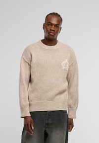 Beige gestrickter Pullover mit Rundhalsausschnitt, gerippten Bündchen und Saum, der ein dezentes cremefarbenes Logo im linken Brustbereich aufweist.