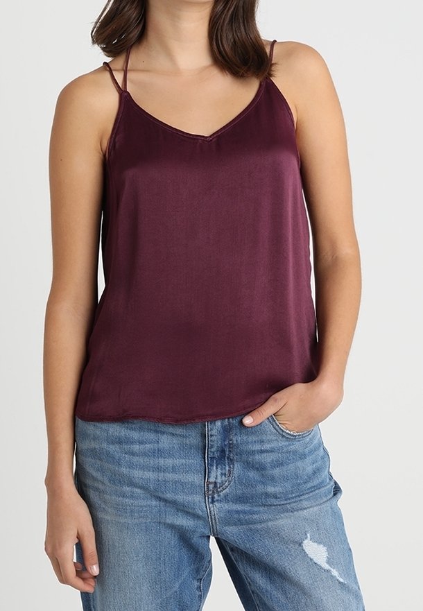 Femme portant un caraco en satin bordeaux sans manches à fines bretelles, associé à un jean bleu clair déchiré.