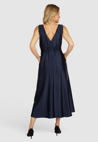 Robe en satin bleu marine avec un dos en V profond, ceinture smockée et jupe fluide. Portée avec des chaussures à talons hauts noires, modèle vu de dos.