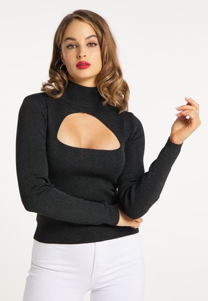 Woman with curled brown hair wearing a black long-sleeve top with chest cutout and white pants, posing against a plain white background.