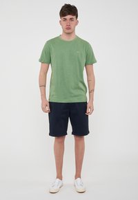 T-shirt verde in cotone con collo rotondo, maniche corte e un piccolo logo sul petto, abbinata a pantaloni corti blu scuro e sneaker bianche.