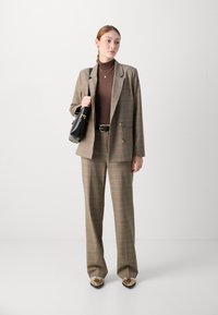 Costume à carreaux marron avec un blazer structuré et un pantalon large, associé à un col roulé marron, un sac noir et des chaussures métalliques.