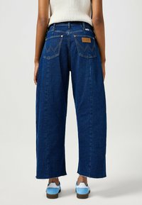 Jeans dżinsowe o szerokich nogawkach, ciemnoniebieskie, z wysokim stanem, z charakterystycznymi tylnymi kieszeniami z wzorem przeszyć oraz postrzępionymi brzegami.