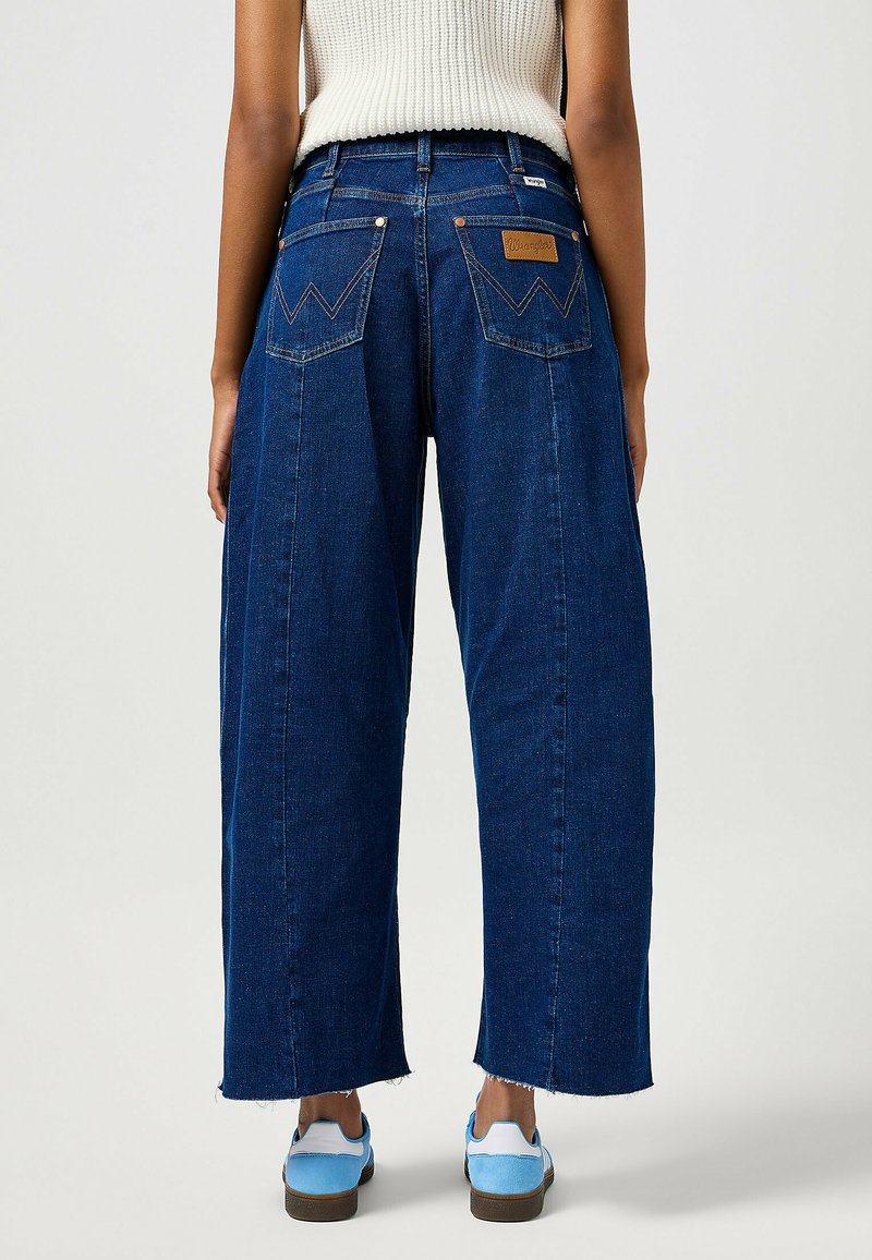 Jean en denim à jambes larges, bleu foncé, taille haute, poches arrière iconiques avec un motif de couture, et ourlets effilochés.