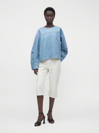 Femme portant un sweat-shirt en denim bleu clair oversize, un short blanc jusqu’au genou et des sandales à talons noirs, debout devant un fond uni.