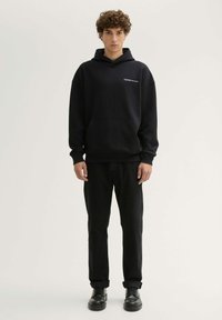 Zwarte hoodie met een kangoeroezak en logo op de borst, gecombineerd met zwarte jeans en zwarte veterschoenen. Zacht, gemêleerd weefsel.