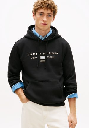 Ung mand iført en sort Tommy Hilfiger hoodie over en blå chambray skjorte og lyse bukser, stående med den ene hånd i lommen.