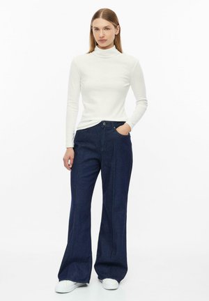 Vrouw staand, gekleed in een witte coltrui, donkerblauwe wijde jeans, witte sneakers, met één hand in de zak tegen een effen witte achtergrond.