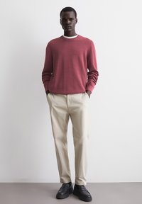 Red knit sweater with a crew neckline, paired with beige trousers and black slip-on shoes. Model stands with hands in pockets against a neutral background.
