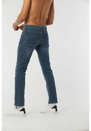 Blauwe spijkerbroek met een slim fit, voorzien van een subtiele S-stiksel op de achterzakken, gedragen met witte sneakers en zwarte accenten.