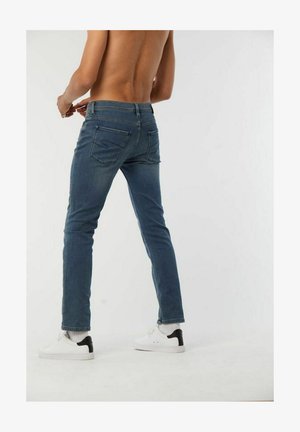 Blauwe spijkerbroek met een slim fit, voorzien van een subtiele S-stiksel op de achterzakken, gedragen met witte sneakers en zwarte accenten.