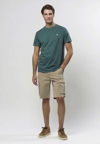 Grönblå crewneck-t-shirt kombinerad med beige cargoshorts med sidofickor. Modellen bär bruna mockasneakers. Enkel, vardaglig design.