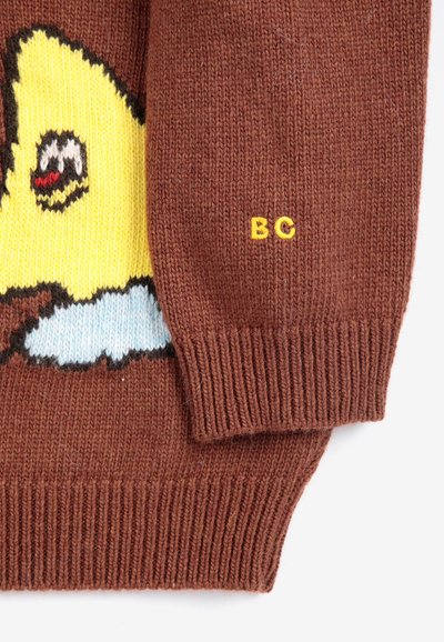Manche de pull tricoté marron avec des lettres jaunes "BC" et une partie d'un dessin d'animal de dessin animé jaune à l'avant.