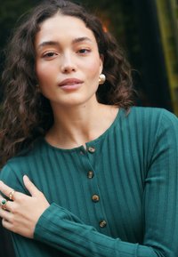 Maglietta a maniche lunghe a coste color verde acqua con scollo rotondo, bottoni sul davanti e tessuto dalla texture morbida. Accessori comprendono orecchini a cerchio dorati e anelli.