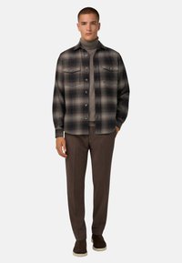 Camisa de franela a cuadros en gris y azul marino, con dos bolsillos en el pecho, cierre de botones, superpuesta a un ligero cuello de tortuga, con pantalones marrones y zapatos oscuros.