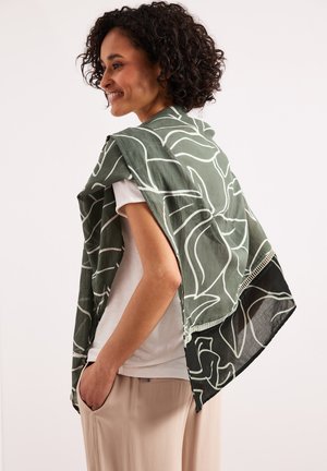 Smiling woman with curly hair drapes green and black patterned scarf over shoulders, wearing white shirt and beige pants with hands in pockets.