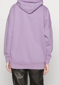 Osoba w dużym, liliowym hoodie oraz czarnych skórzanych spodniach, pokazana od tyłu na jednolitym jasnym tle.