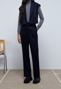 Paket tuksedo bergaris navy dengan blazer tanpa lengan yang dipangkas dan celana lebar, dipadukan dengan turtleneck lengan panjang abu-abu dan alas kaki hitam.