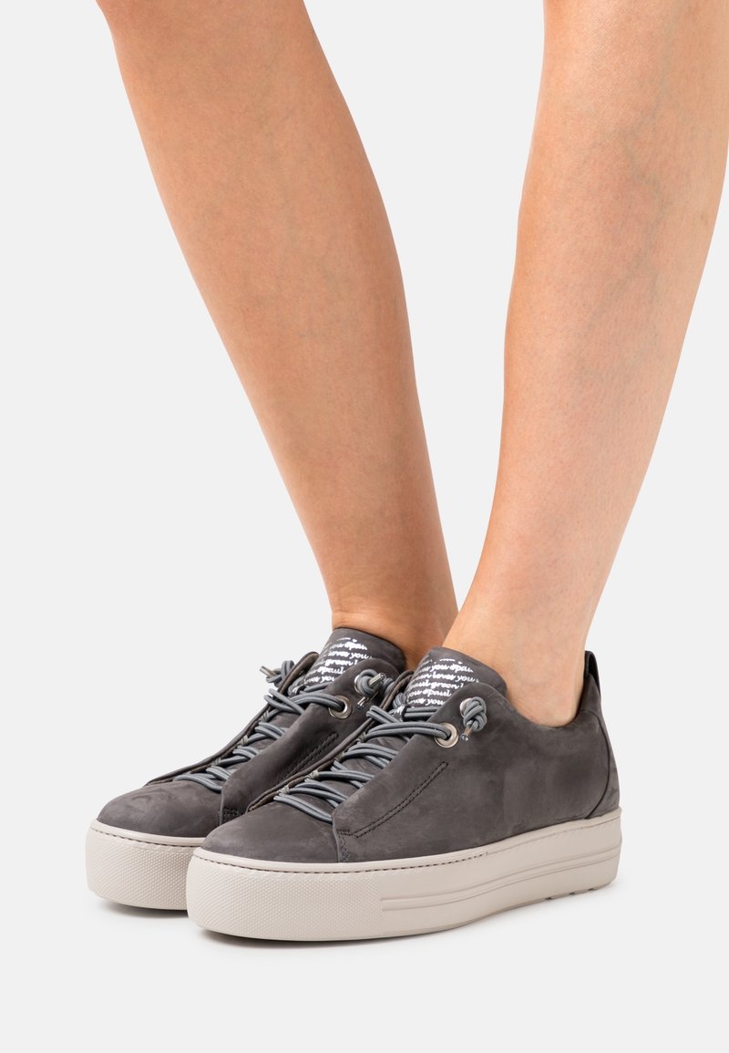 Jambes portant des baskets compensées en suède gris avec des semelles épaisses blanches et des patchs à motifs sur la languette, montrées sur un fond uni.