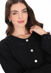 Schwarzer strukturierter Cardigan mit Rundhalsausschnitt, verziert mit silbernen Knöpfen und einem Perlenakzent am Ausschnitt. Weicher Stoff, entspannte Passform.