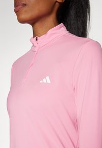 Jasnoróżowa sportowa bluzka z półzamkiem, o gładkiej fakturze i dopasowanym kroju, z białym logo marki na lewej stronie klatki piersiowej.