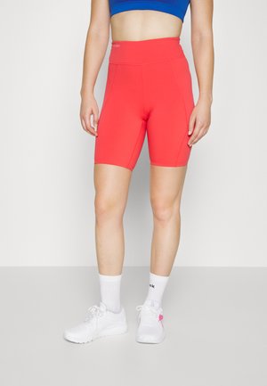 Κόκκινα ποδηλατικά σορτς με ψηλή μέση και λείο, ελαστικό ύφασμα. Συνδυασμένα με λευκά αθλητικά παπούτσια και κάλτσες, συνοδευόμενα από μπλε κοντομάνικη μπλούζα.