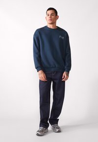Sudadera azul marino con cuello redondo y logotipo bordado, combinada con jeans azul oscuro y zapatillas grises con acentos de malla.