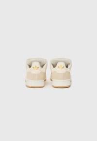 Sneakers de camurça bege com sola de borracha branca, apresentando detalhes em branco e dourado no calcanhar e no logotipo, com uma forma slim e arredondada.